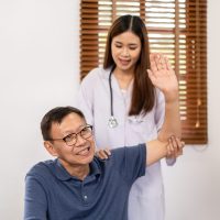 Female physical therapist working and doing arm rehabilitation therapy for elderly patient sitting on hospital bed and talking to giving advice on treatment to patient.